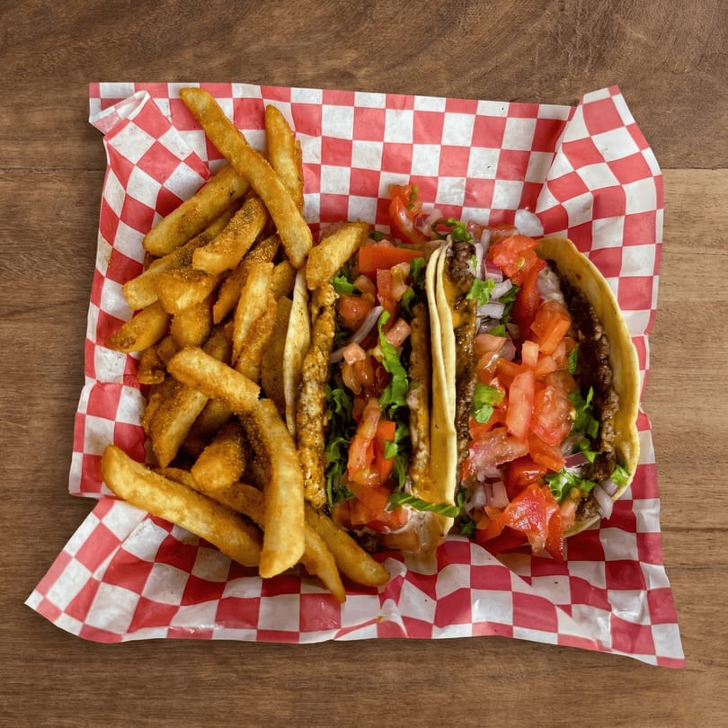Two tacos with ground beef, lettuce, tomato, and onion, served with seasoned fries on checkered paper.