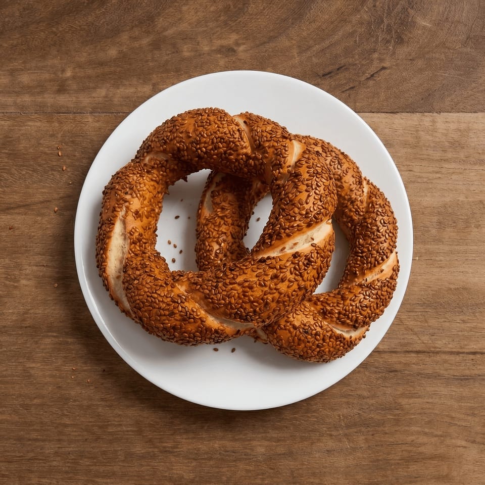 A golden-brown, sesame-covered simit, a Turkish bagel, on a white plate atop a wooden table.