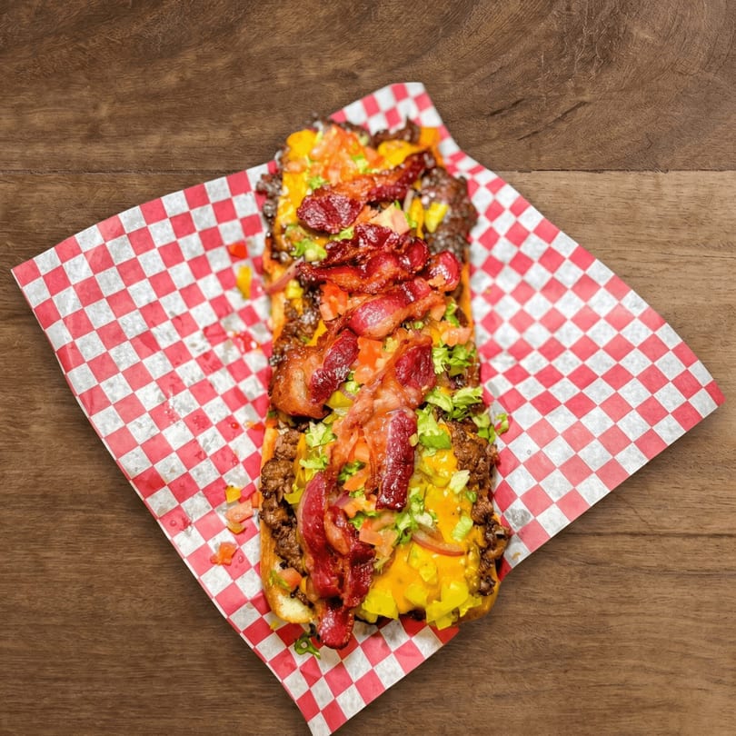 Loaded hot dog with bacon, cheese, and vegetables on red and white checkered paper.