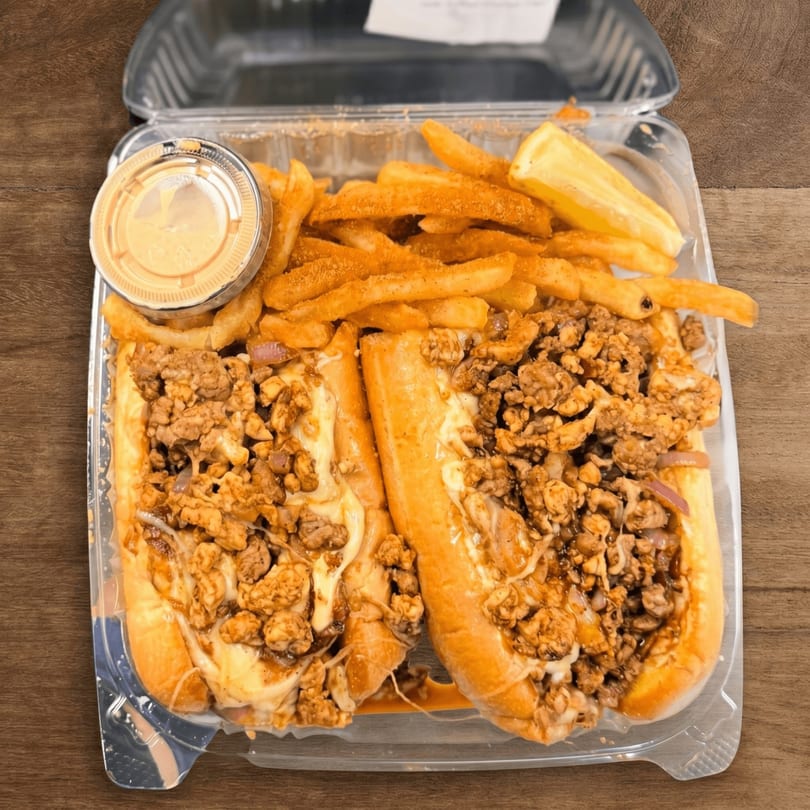 Two cheesesteak sandwiches with fries and dipping sauce in a clear to-go container.