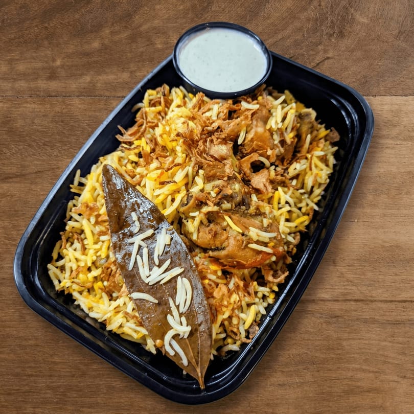 Chicken biryani with fried onions and a bay leaf, served with a side of raita.