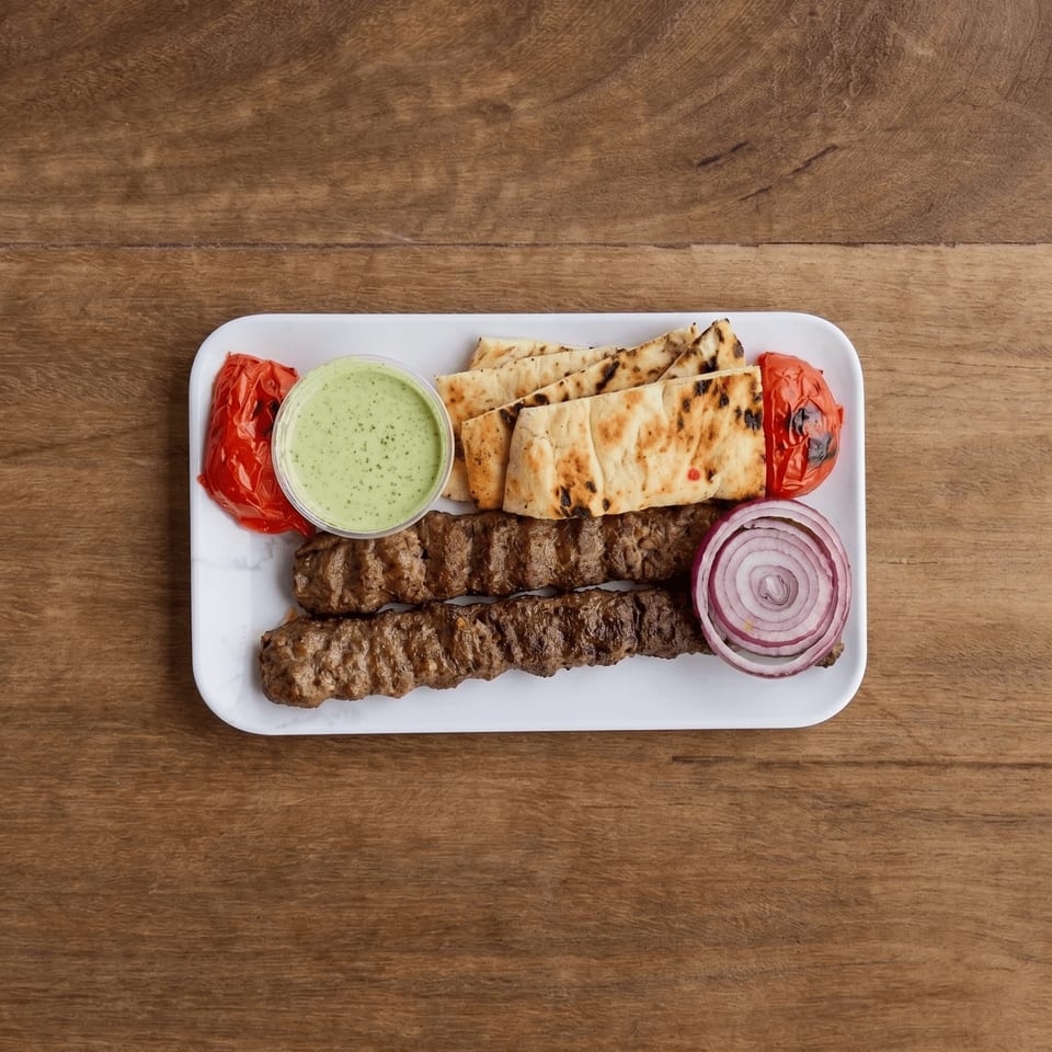 Two grilled meat skewers, flatbread, roasted tomatoes, red onion, and green sauce on a white plate.