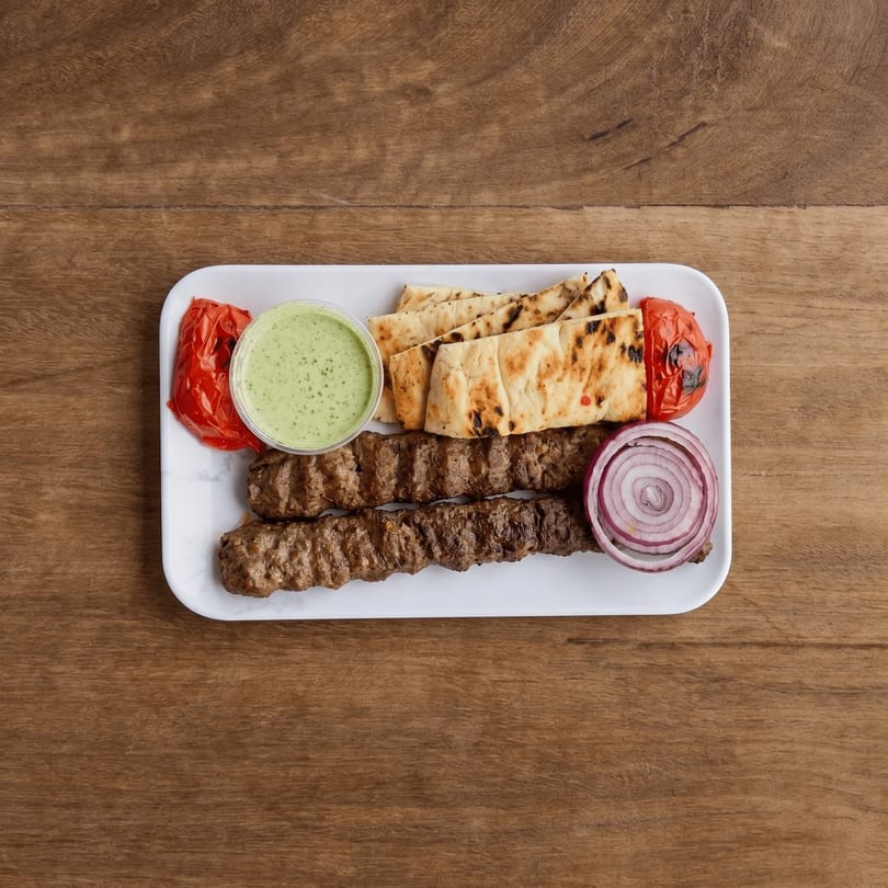 Two grilled meat skewers, flatbread, roasted tomatoes, red onion, and green sauce on a white plate.