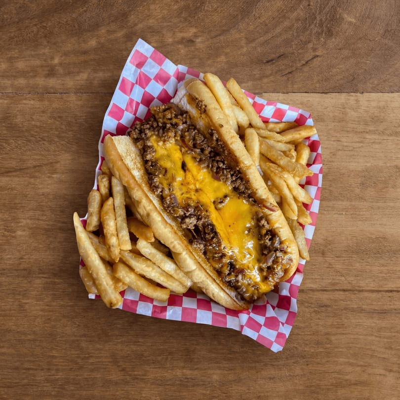 Cheesesteak sandwich with melted cheese and fries in a red and white checkered basket on a wooden table.
